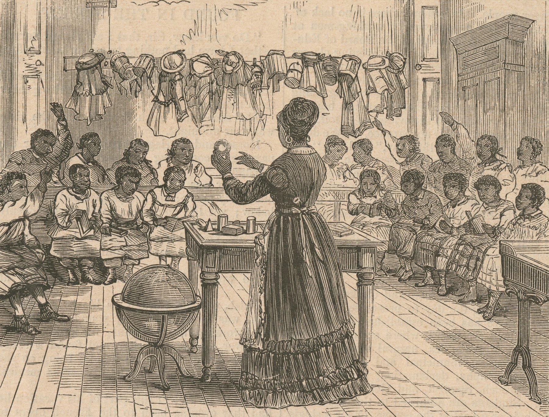 Classroom with students against the far wall and teacher standing in foreground with desk and globe.
