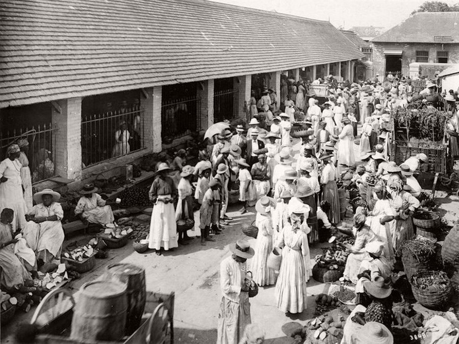 Market with sellers seated before baskets, and many individuals in white clothing milling about.