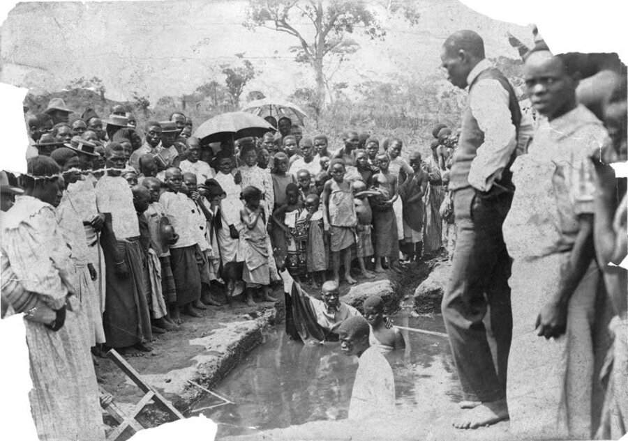 Boy and man with arm raised standing in water surrounded by crowd of onlookers, some with umbrellas.