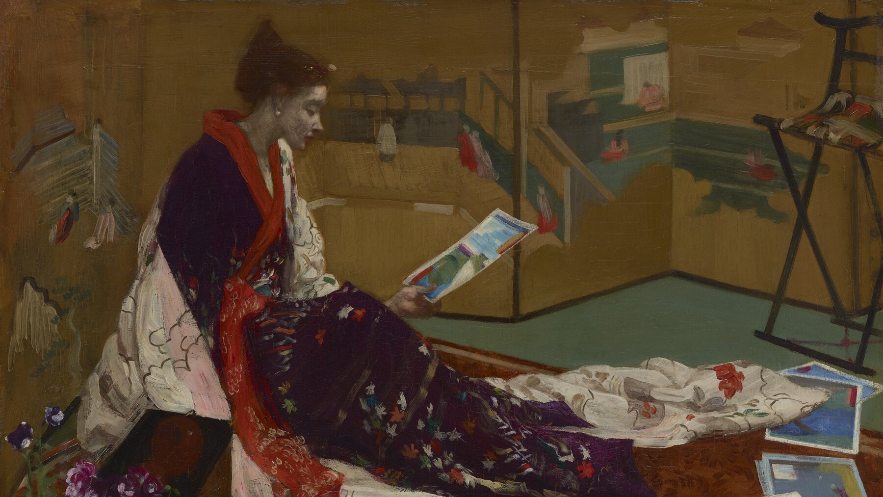 Caprice in traditional Japansese dress, seated and examining one of several illustrations, with gold screen in background.