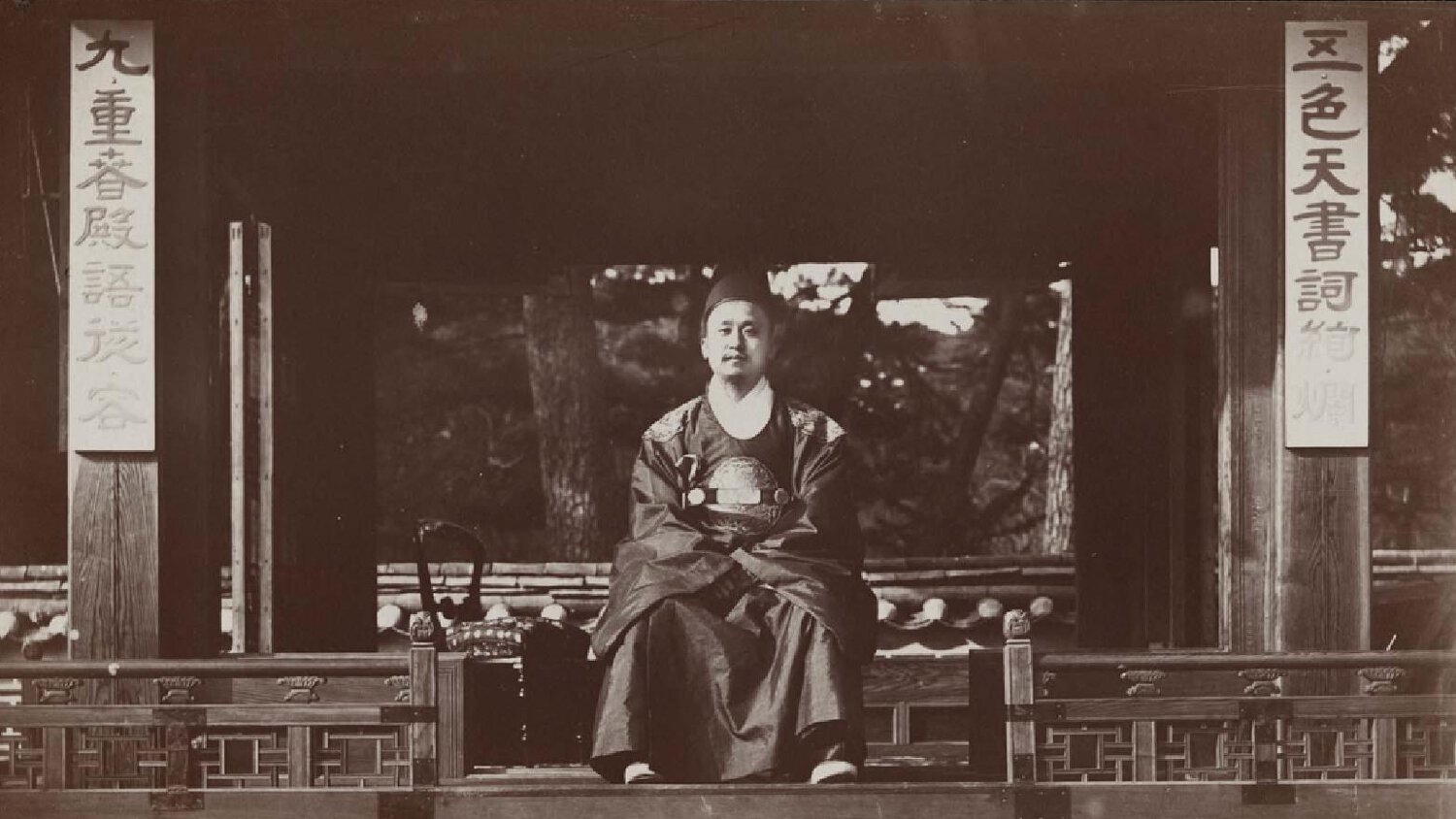 King of Korea seated, facing forward with arms across chest, pillars of Korean writing on each side.