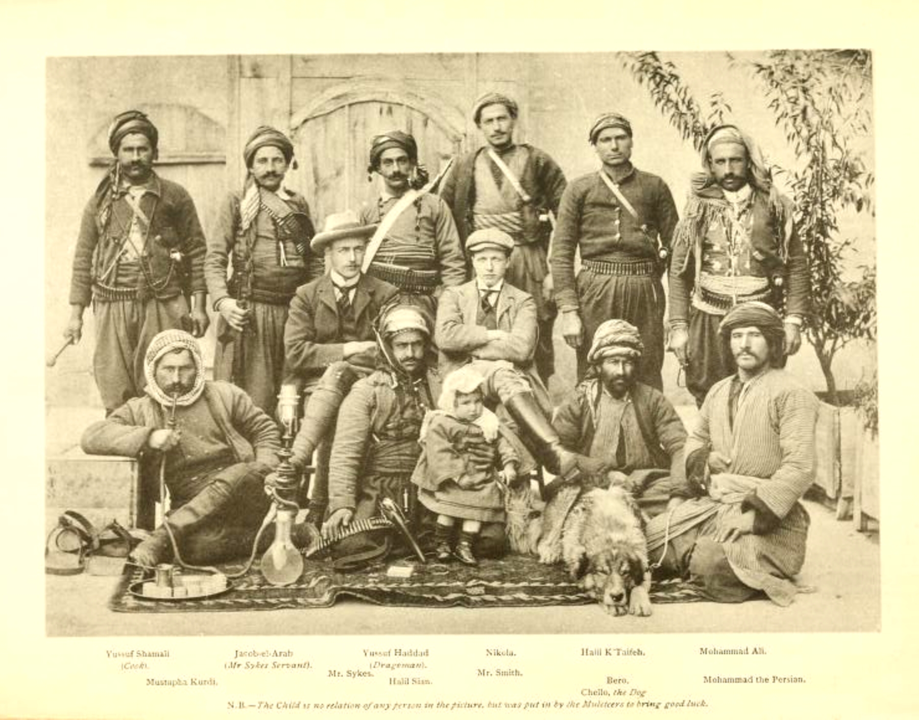A photograph depicting twelve men wearing head coverings, posed with a child, dog, and other supplies
