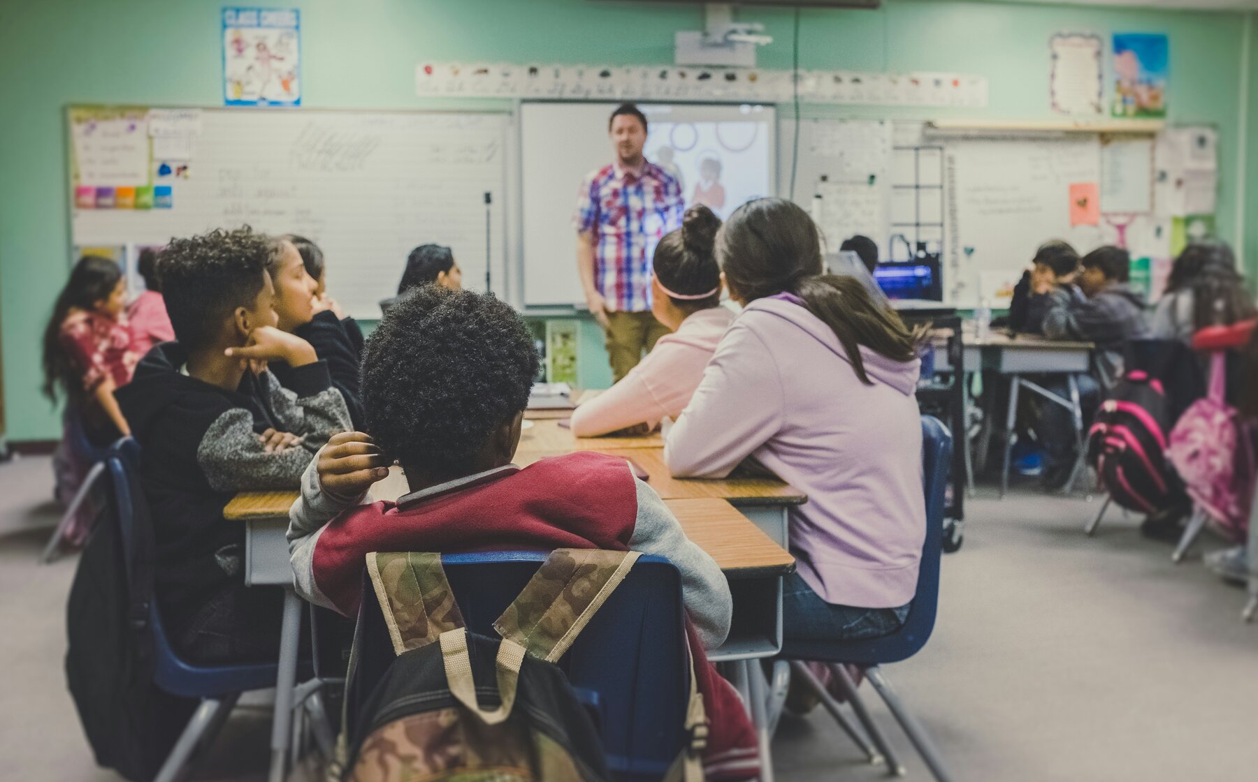 View of classroom from back, children's heads in foreground, stading teaching in distance.