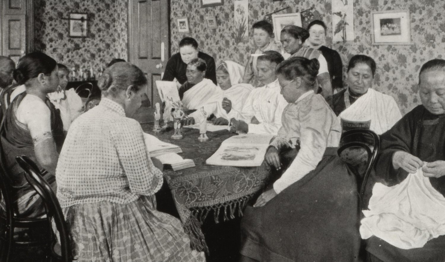 Interior scene with a large group of women seated around a table, many looking at documents.