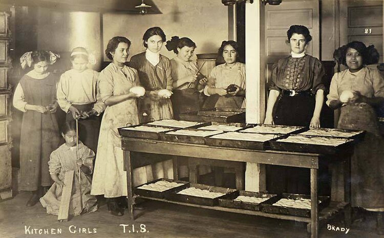 A group of seven women, randing in age from adolescent to adult, gathered around a kitchen table.